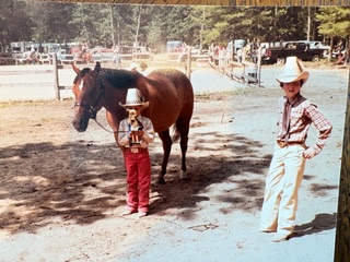 A young girl in red and a cowboy hat holding up a trophy in front of a brown horse. To the right, a young boy in white chaps and a white hat.